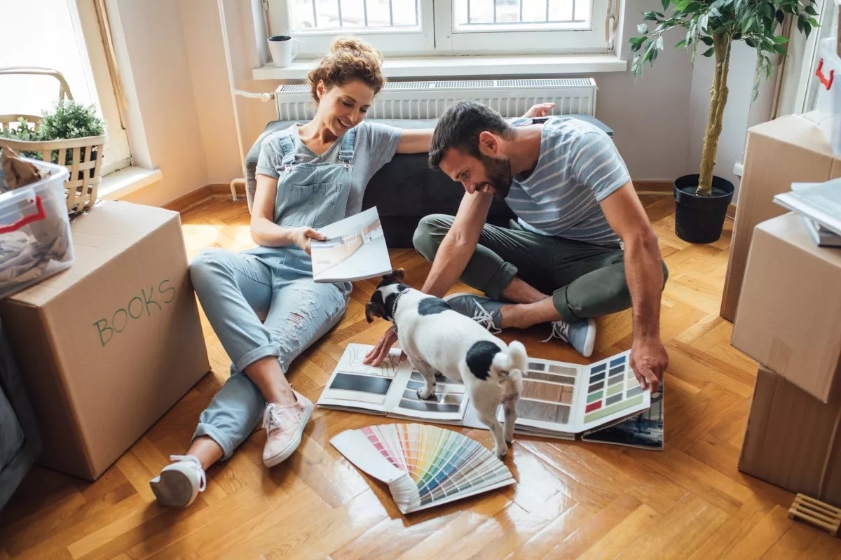 A couple sitting on the floor choosing hardwood styles and colors. There's a small dog interrupting them by standing on their materials. The couple is joyous.